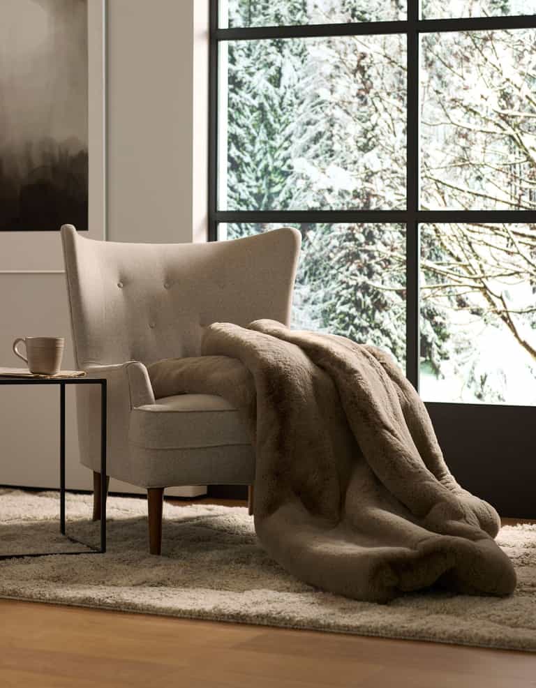 A cozy beige armchair with a Cozy Earth Cuddle Blanket draped over it sits beside a small table with a mug, in front of a large window overlooking snowy trees.|Color:Walnut