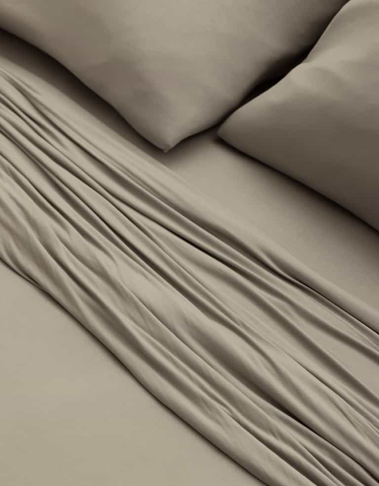 A close-up of a bed made with Cozy Earth’s Bamboo Jersey Sheet Set in taupe, featuring two matching pillows and showcasing the soft fabric folds and smooth texture.|Color:Driftwood
