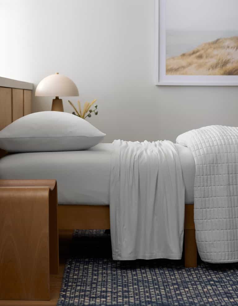 A bed made with Cozy Earth’s Bamboo Jersey Sheet Set rests against a wooden headboard. A wooden bench stands at the foot, while a lamp, small vase, and framed landscape photo accentuate the minimal, serene bedroom.|Color:White