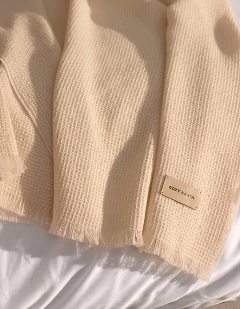A Cozy Earth Alpaca Waffle Throw in creme, featuring a textured waffle weave and fringed edges, is draped over a white surface. A beige tag on the throw displays the brand name "Cozy Earth.|Color:Creme
