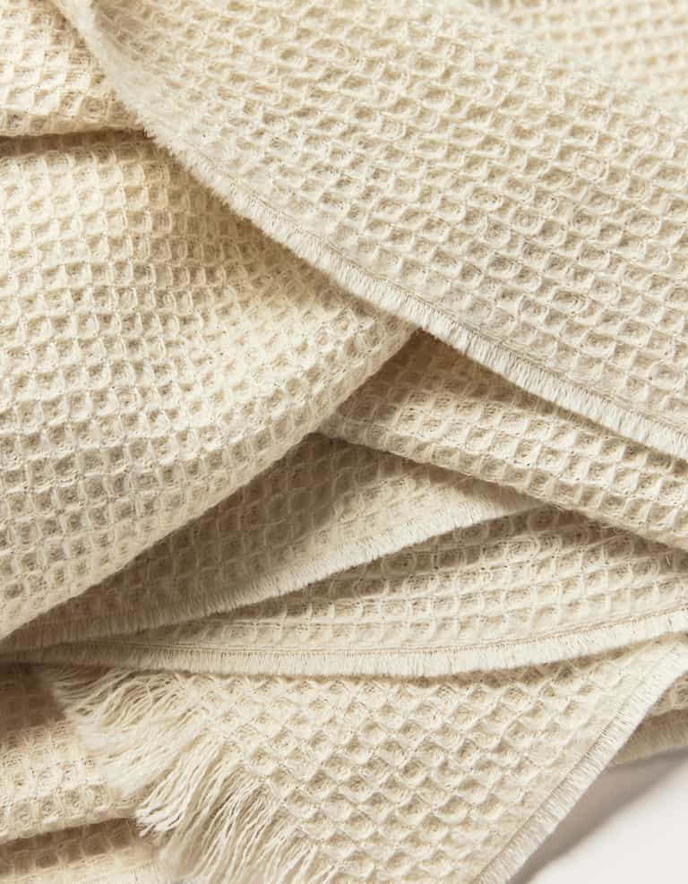 A close-up of the Cozy Earth Alpaca Waffle Throw highlights its cream-colored waffle weave texture and fringed edges, showcasing its soft and cozy appearance.|Color:Creme