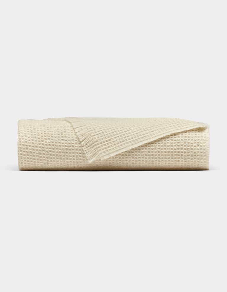 The Cozy Earth Alpaca Waffle Throw, in beige with a fringed edge, is neatly folded and displayed against a plain light gray background.|Color:Creme