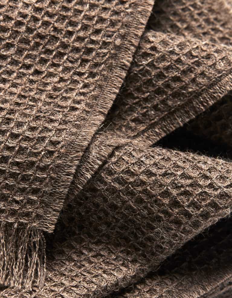 Close-up of the Alpaca Waffle Throw by Cozy Earth, highlighting its brown waffle weave texture, soft fibers, and layered folds. Fringed edges are visible in the lower left corner, emphasizing its cozy appeal. |Color:Ebony