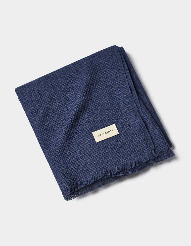A folded Alpaca Waffle Throw from Cozy Earth in dark blue, featuring a textured pattern, fringed edges, and a small rectangular "Cozy Earth" label sewn near one corner.|Color:Slate Blue
