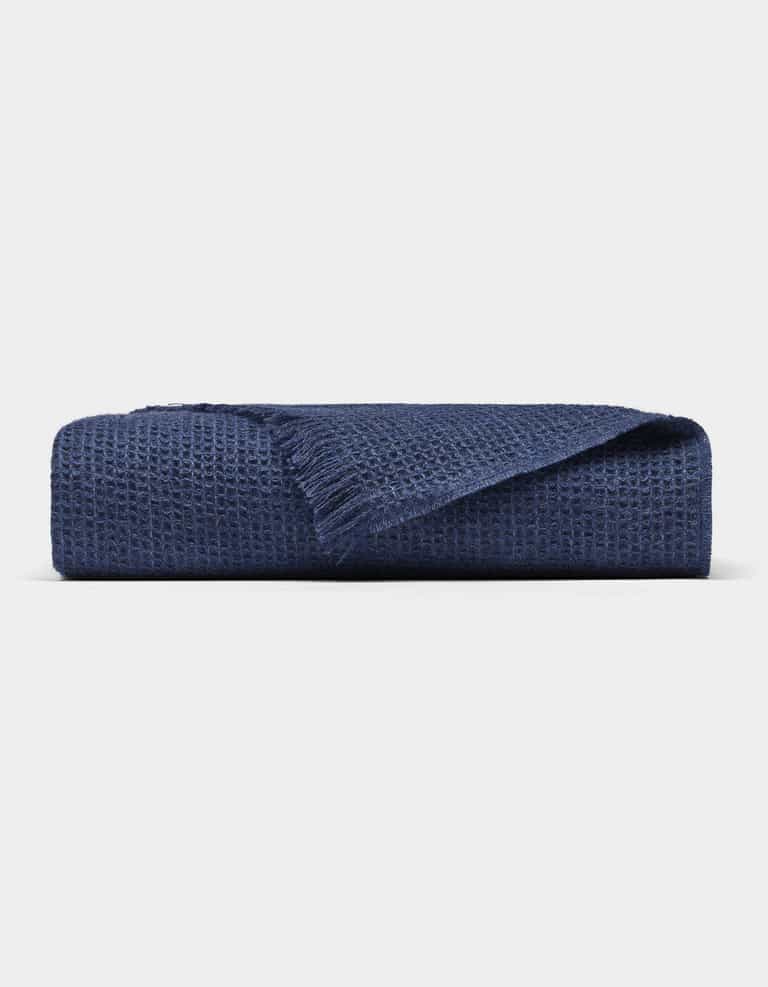 The Cozy Earth Alpaca Waffle Throw, in navy blue with a waffle texture and fringed edge, is shown folded against a plain light background.|Color:Slate Blue