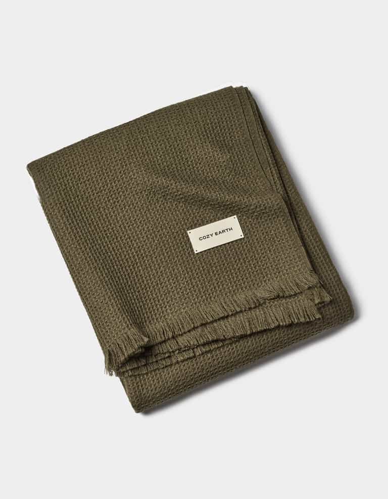 A folded olive green Alpaca Waffle Throw by Cozy Earth features a waffle texture, fringed edges, and a white tag with the brand name, set against a plain white background.|Color:Utility Green