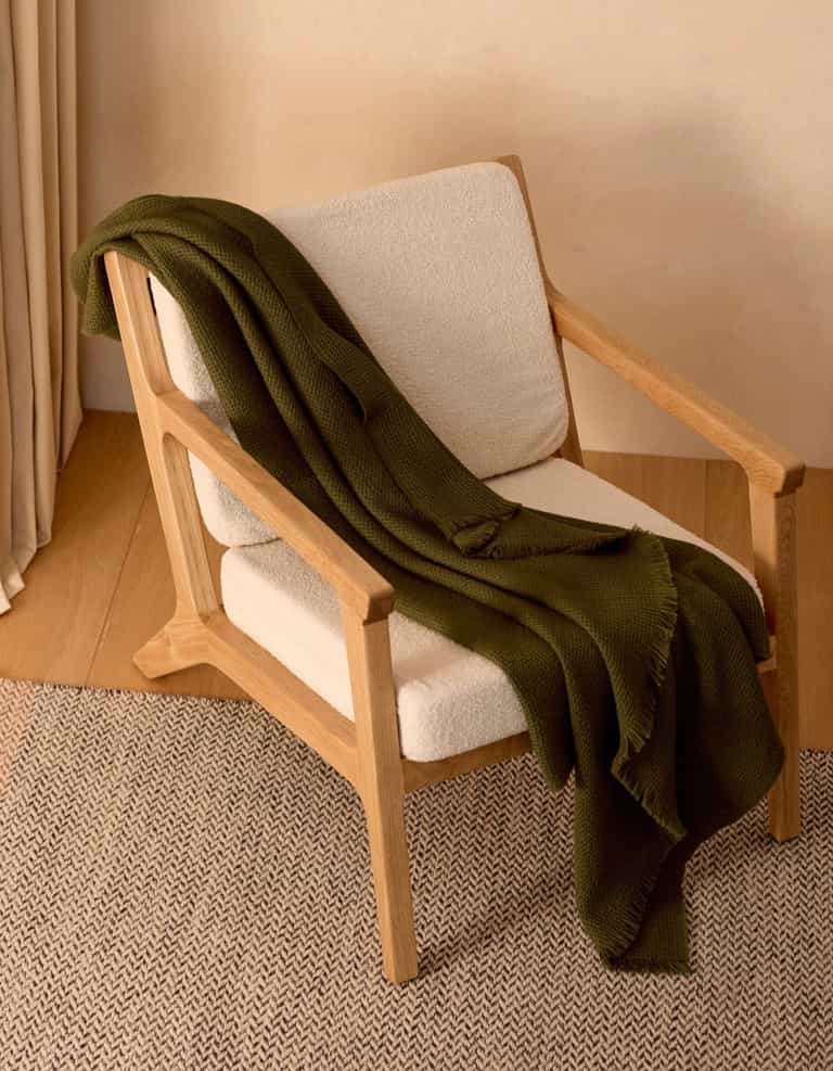 A wooden armchair with cream-colored cushions sits on a woven rug. A Cozy Earth Alpaca Waffle Throw in dark green is draped over the chair, adding a cozy and inviting touch.|Color:Utility Green