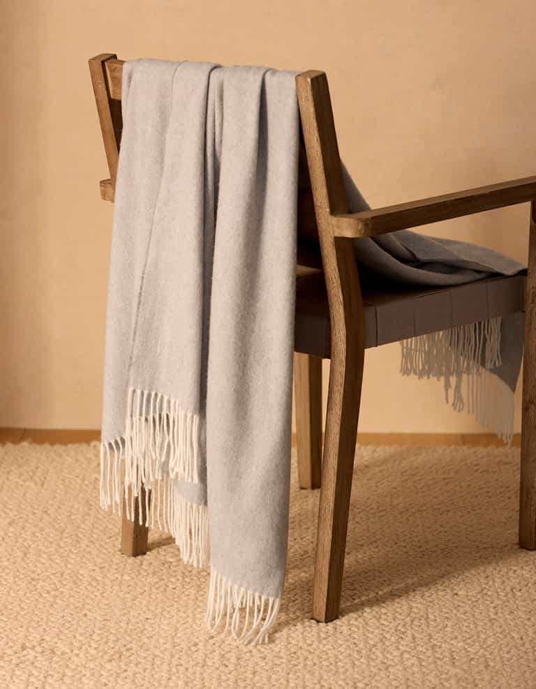 The Bamboo Herringbone Tassel Throw by Cozy Earth, in light gray with fringe, is draped over a cushioned wooden chair on a woven beige rug against a neutral wall.|Color:Cloud