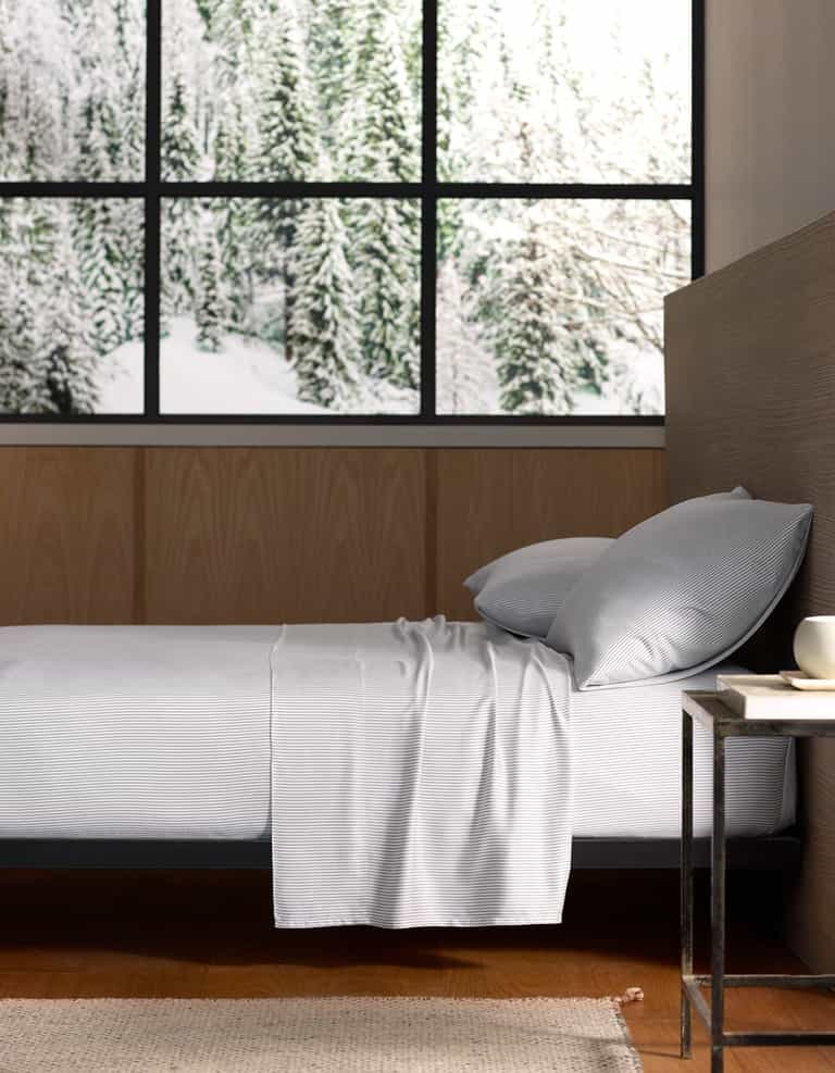 A bed with Cozy Earth Bamboo Flat Sheets and pillows is beside a small table with a cup. Large windows reveal a snowy forest, filling the room with natural light.|Color:Charcoal Pinstripe