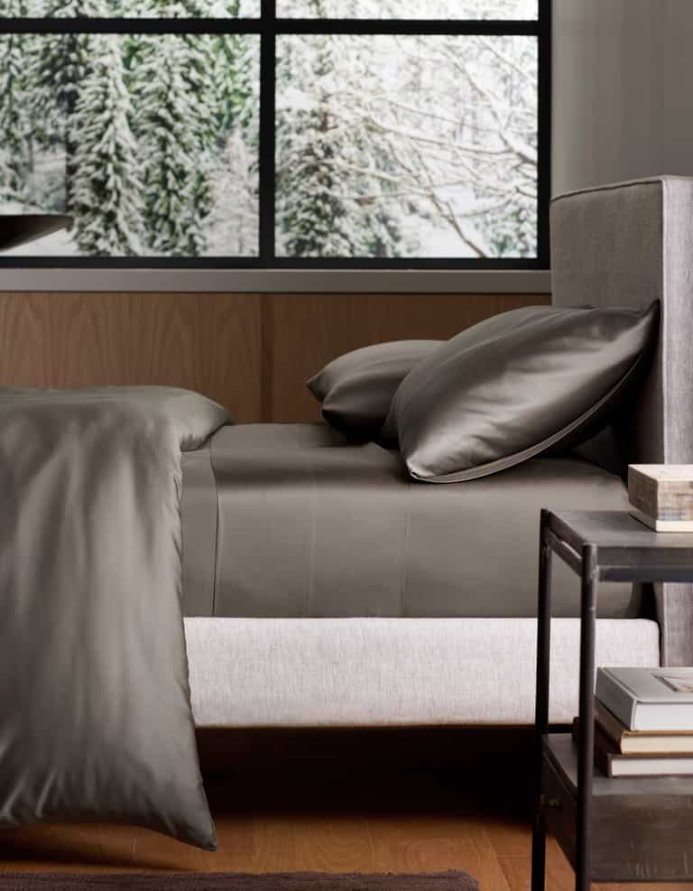 A neatly made bed with Cozy Earth’s Bamboo Sheet Set in dark gray rests by a modern nightstand. Behind the bed, a large window reveals a snowy forest with pine trees. |Color:Charcoal