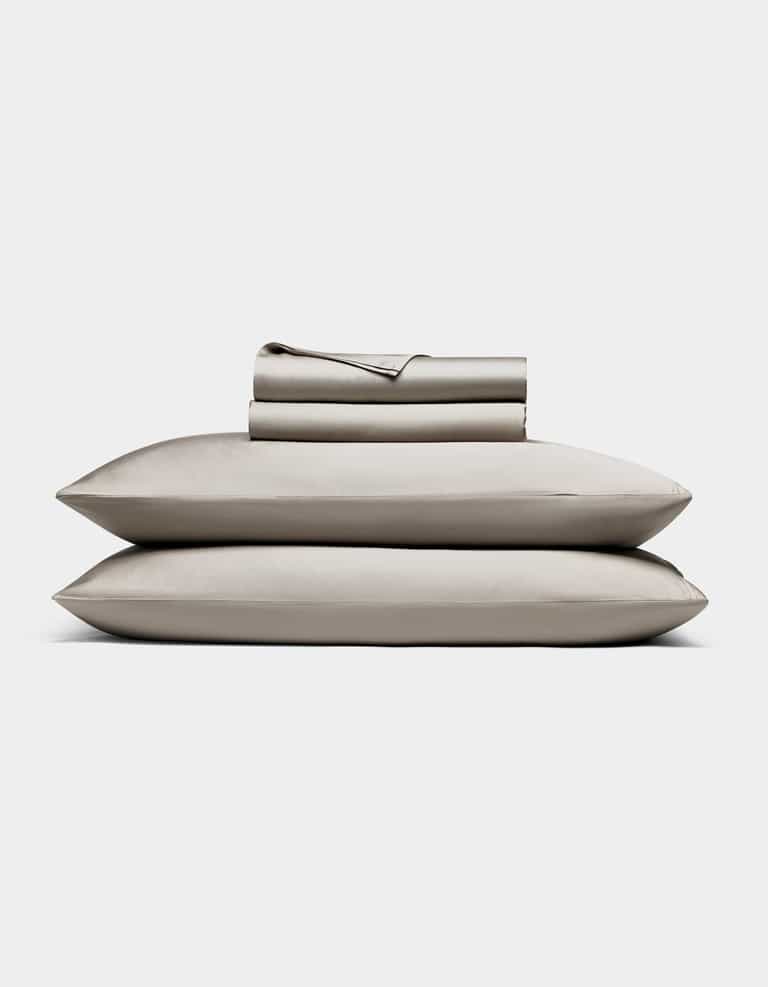 The Cozy Earth Bamboo Sheet Set, in light gray, includes neatly folded bed sheets and pillowcases stacked atop two pillows, all shown against a plain white background.|Color:Dove Grey