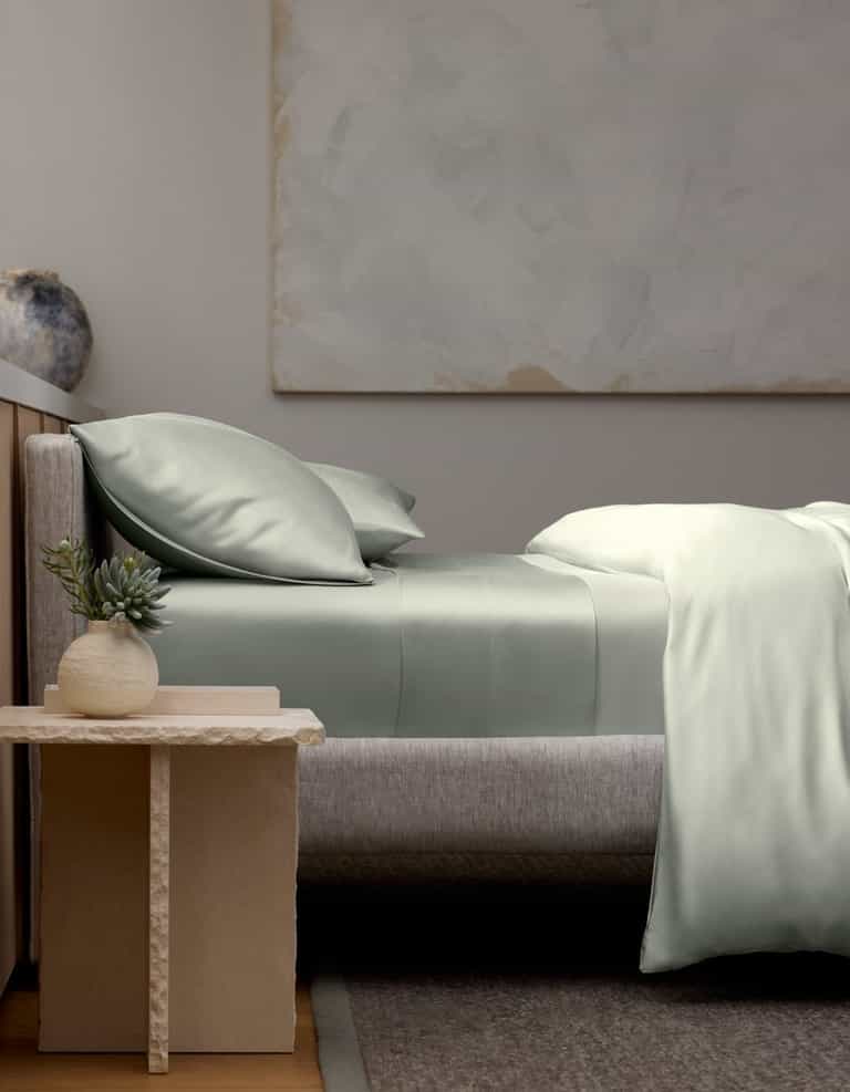 A modern bedroom featuring a neatly made bed with Cozy Earth’s Bamboo Fitted Sheet, two pillows, and a gray upholstered headboard. A small table with a potted succulent and ceramic vase sits beside the bed. Abstract art hangs on the wall.|Color:Laurel