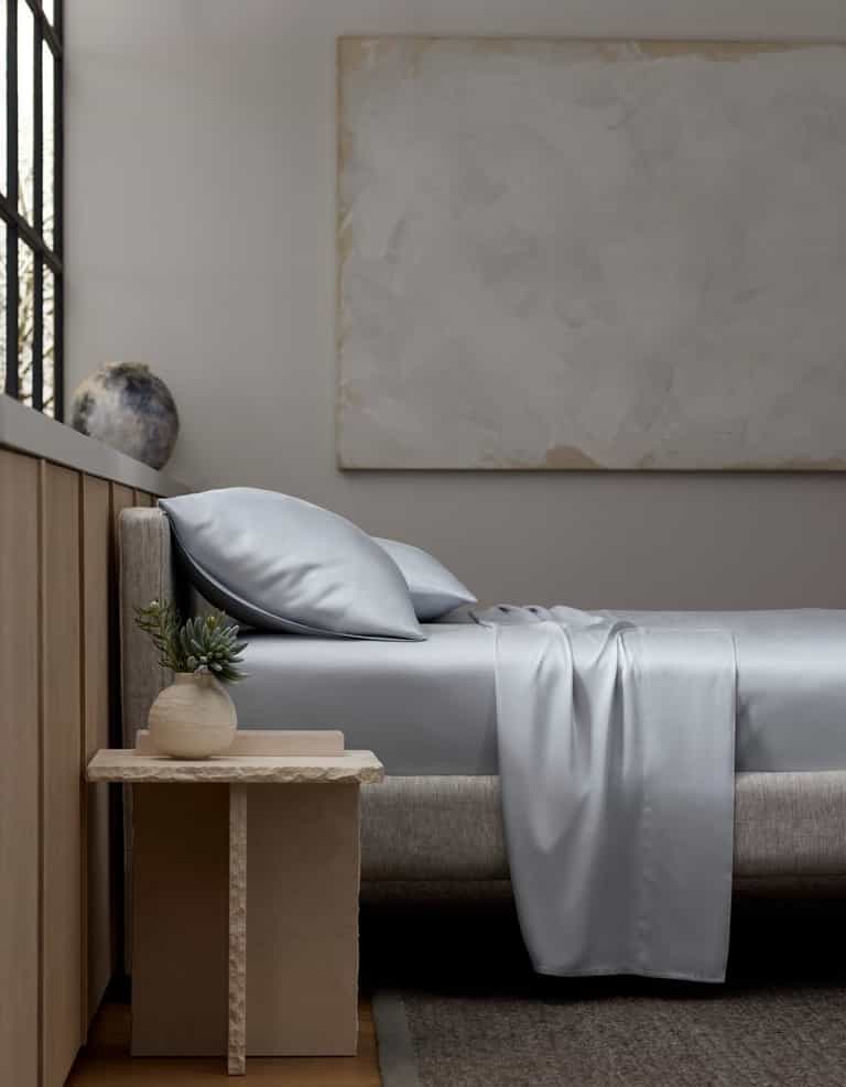 A minimalist bedroom with a light gray upholstered bed, Cozy Earth Bamboo Sheet Set in silver-gray, two pillows, a beige nightstand with a small potted plant, a large abstract painting, and a window letting in natural light.|Color:Shore