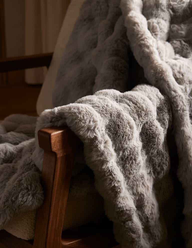 The Bubble Cuddle Blanket by Cozy Earth, with its soft gray faux fur, is draped over a wooden chair, adding a cozy and inviting touch to the space.|Color:Light Grey