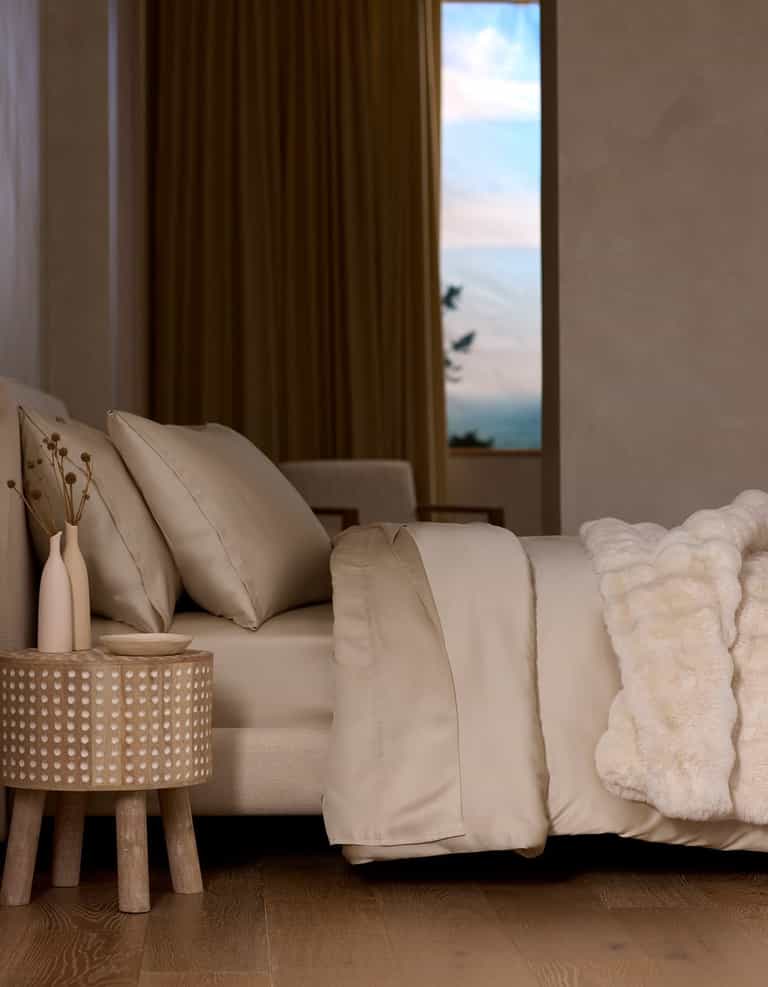 A neatly made bed features the Cozy Earth Bamboo Duvet Cover in beige and a white faux fur blanket, beside a textured wooden nightstand with two vases. Large curtained windows reveal a scenic sky.|Color:Driftwood