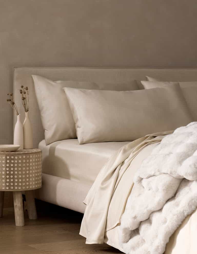 A neatly made bed features Cozy Earth Bamboo Pillowcases in cream, a soft white blanket, and a nightstand with minimal vases of dried plants against a beige wall.|Color:Oat