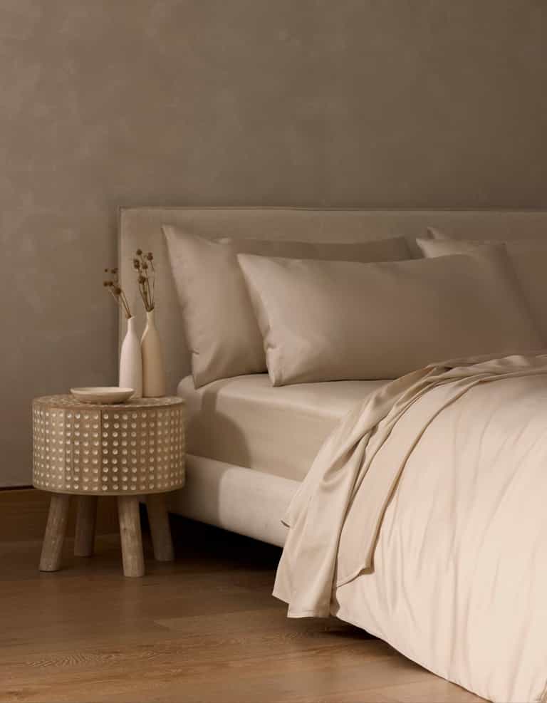 A minimal beige bedroom showcases a neatly made bed with smooth, light Bamboo Sheet Set by Cozy Earth. A small round side table with textured pattern holds two simple vases with dried flowers. The space features wooden flooring and neutral walls.|Color:Driftwood