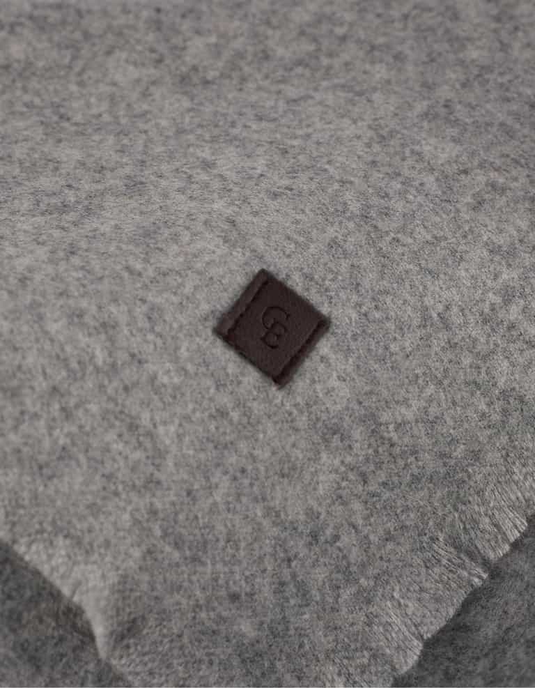 A close-up of Cozy Earth's Cashmere Fringe Blanket shows its soft, light gray fabric accented with a small, dark brown square patch embossed with a "B.|Color:Pebble