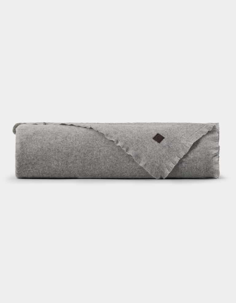 A neatly folded light gray Cozy Earth Cashmere Fringe Blanket with a small dark brown patch on one corner, displayed against a plain white background.|Color:Pebble