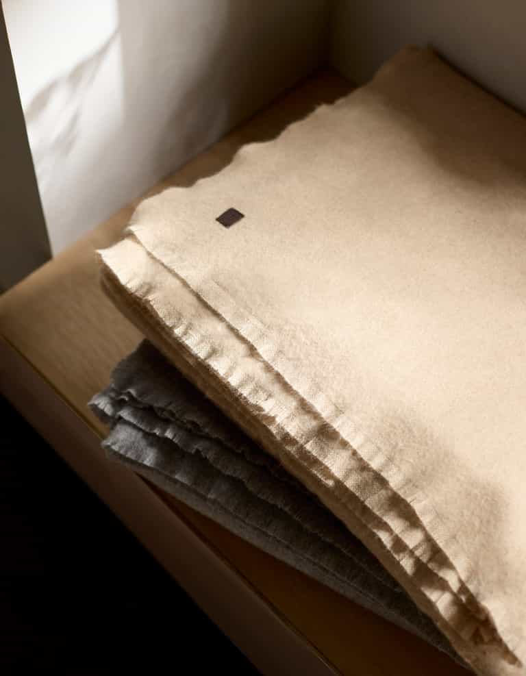 A Cozy Earth Cashmere Fringe Blanket in beige is neatly folded atop a gray one on a wooden surface, with soft light casting gentle shadows around them.|Color:Dune