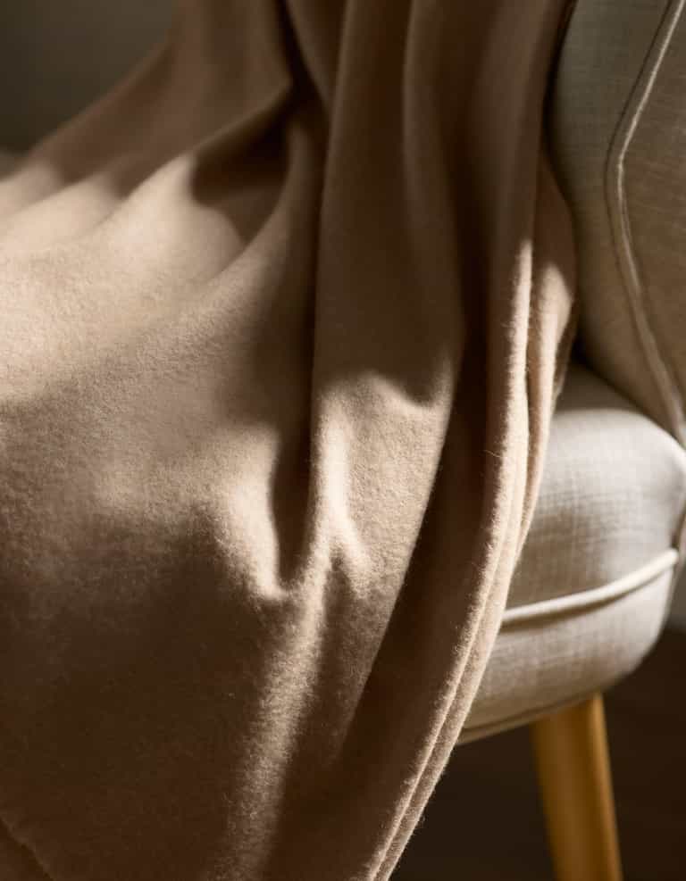 The Cashmere Fringe Blanket by Cozy Earth, in soft light brown, is draped over a light beige upholstered chair with wooden legs and bathed in natural sunlight.|Color:Dune
