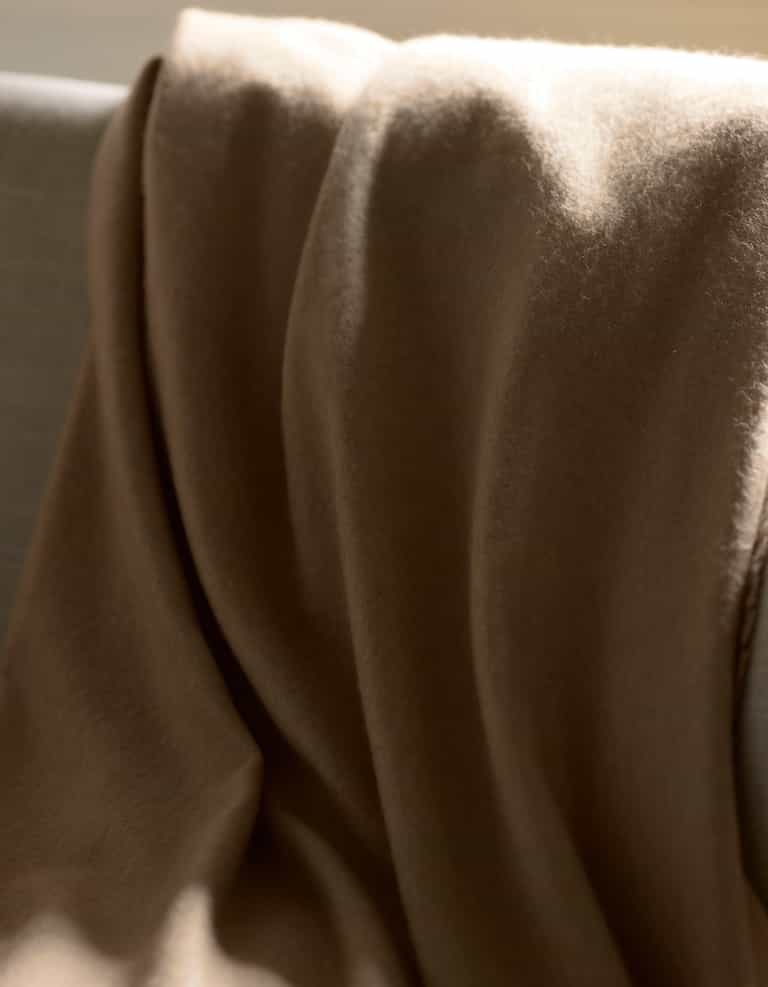 The Cozy Earth Cashmere Fringe Blanket, in a soft brown shade, is draped over a light-colored sofa as sunlight casts gentle highlights and shadows on its luxurious fabric.|Color:Dune
