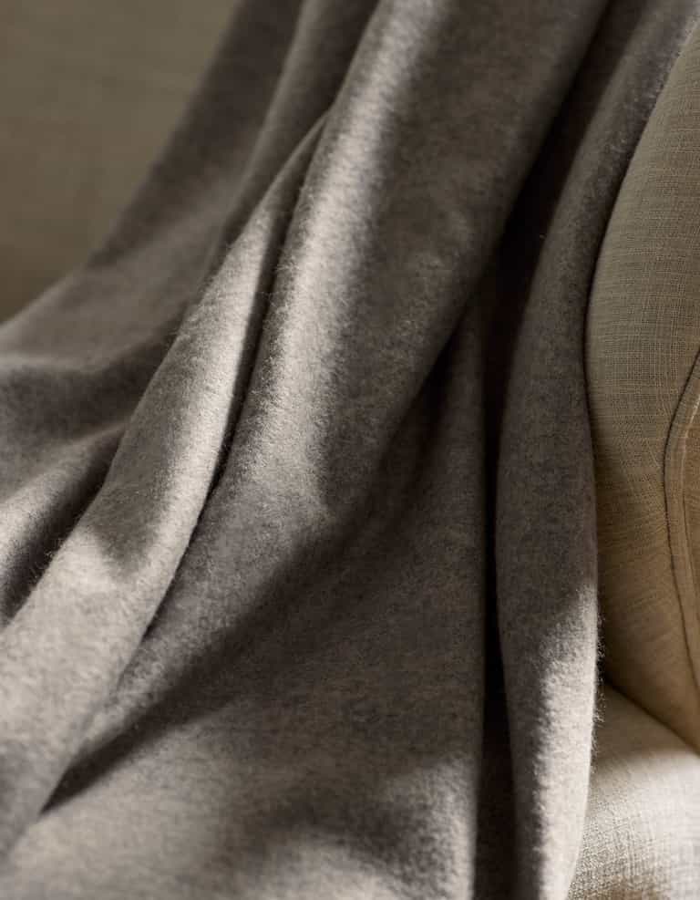 A close-up of the Cozy Earth Cashmere Fringe Blanket, featuring a soft, gray fabric with a cozy, slightly fuzzy texture and gentle folds, draped over the arm of a light beige upholstered chair.|Color:Pebble