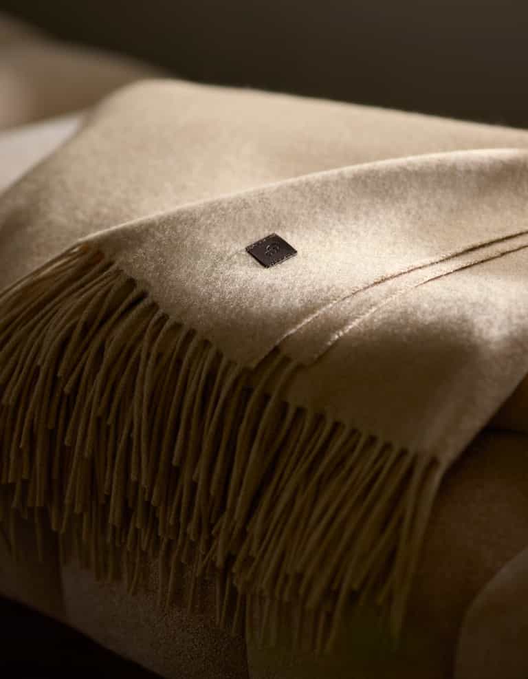 A close-up of the Cozy Earth Cashmere Tassel Throw, a beige cashmere blanket with fringe and a small square leather label, softly lit by natural light.|Color:Dune