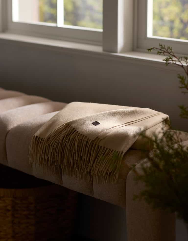 A Cozy Earth Cashmere Tassel Throw in beige is neatly folded on a cushioned bench near a window, softly lit by natural light with greenery partially visible in the foreground.|Color:Dune