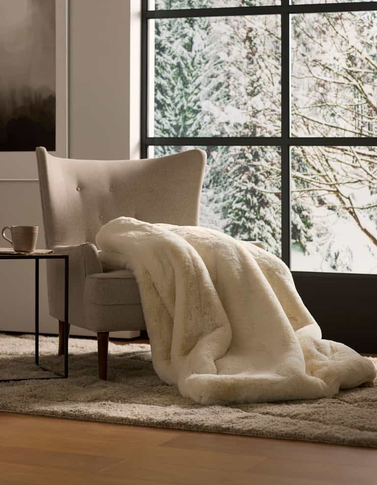 A beige armchair with a Cozy Earth Cuddle Blanket is placed by a small table with a mug, set before large windows overlooking a snowy, tree-filled landscape.|Color:Creme