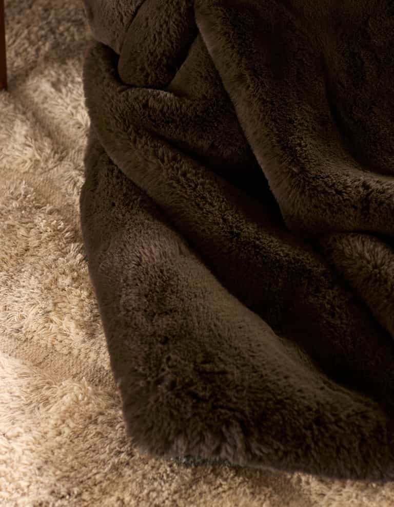 A close-up of the Cozy Earth Cuddle Blanket in soft, dark brown faux fur draped over a light beige shaggy carpet, showing off the distinct textures of both the blanket and carpet.|Color:Espresso