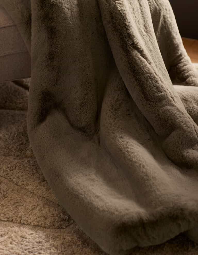 The Cozy Earth Cuddle Blanket, a soft plush brown faux fur throw, is draped over a couch edge and rests on a textured beige carpet in a warmly lit, cozy room.|Color:Walnut