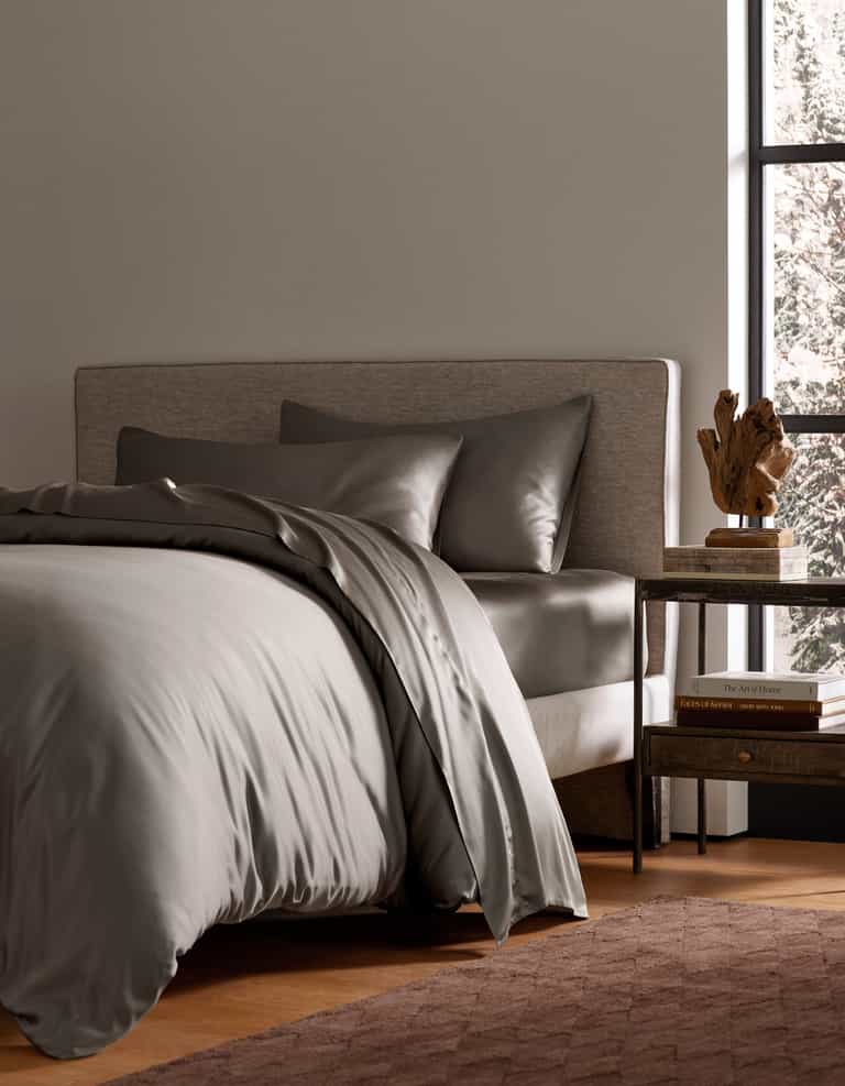 A neatly made bed features the Bamboo Bedding Deluxe Bundle by Cozy Earth in gray, two matching pillows, a light upholstered headboard, a wooden nightstand with books and a sculpture, and natural light streaming through a large window.|Color:Charcoal