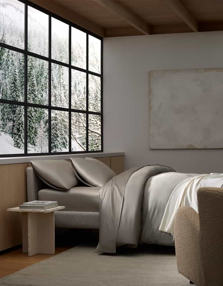 A cozy bedroom featuring Cozy Earth's Bamboo Bedding Deluxe Bundle, a gray bed and chair by the window with snowy forest views. Neutral tones, a soft rug, and a minimalistic painting create a serene, modern vibe.|Color:Dove Grey