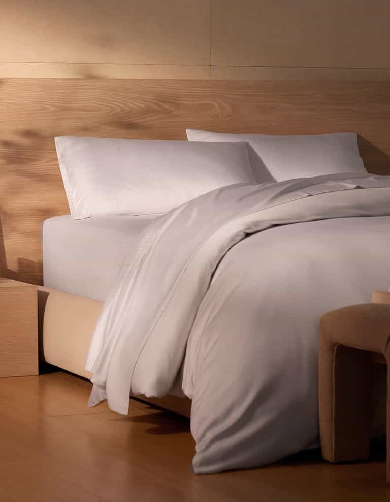 A neatly made bed features Cozy Earth's Bamboo Sheet Set in white, paired with pillows, a wooden headboard, light wood floors, and a beige upholstered bench in a softly lit minimalist bedroom.|Color:Driftwood Pinstripe