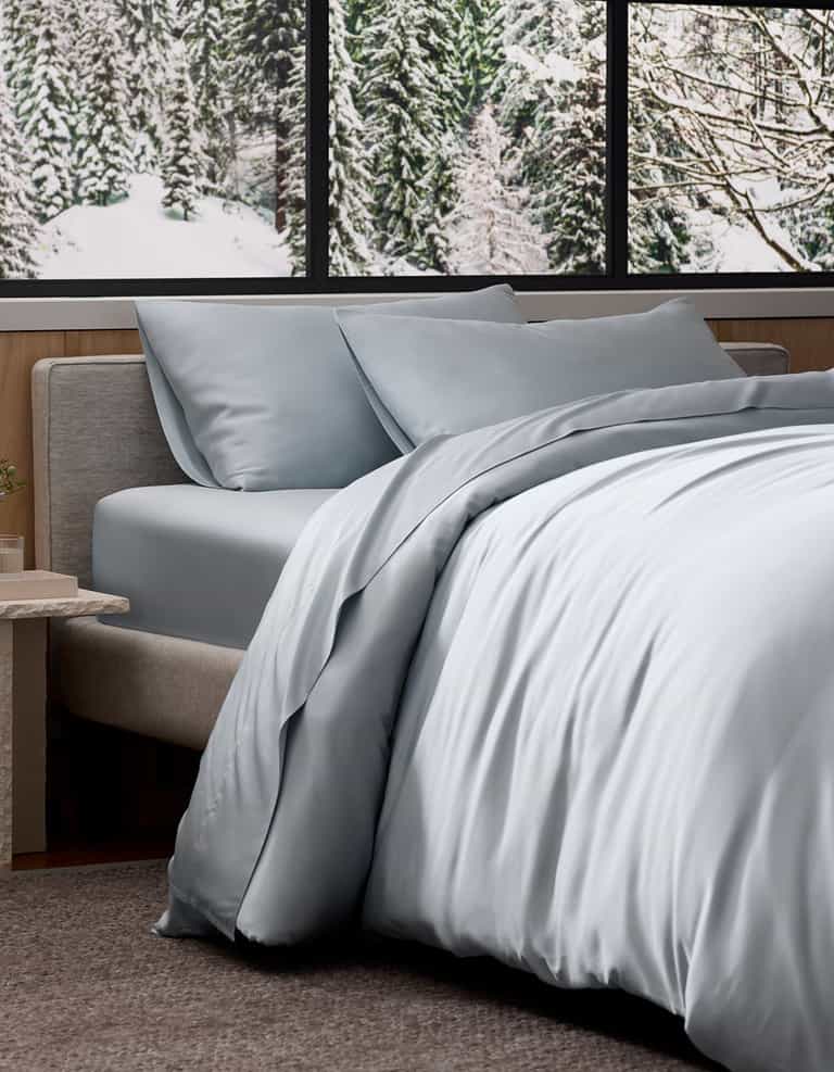 A neatly made bed featuring Cozy Earth's Bamboo Bedding Deluxe Bundle in light gray sits by a window overlooking snowy trees, creating a cozy, modern ambiance.|Color:Shore