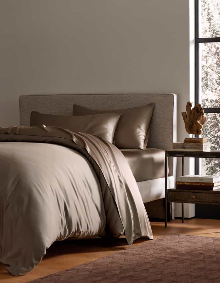 A modern bedroom features a neatly made bed with Cozy Earth’s Bamboo Bedding Deluxe Bundle in taupe, paired with pillows. A wooden nightstand nearby holds books, a lamp, and decor, as sunlight adds warmth to the inviting space.|Color:Walnut