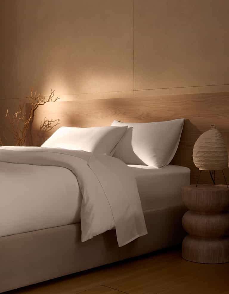 A neatly made bed featuring Cozy Earth's Bamboo Duvet Cover is set beside a wooden nightstand with a round lamp; beige walls, natural wood accents, and a dried branch in the corner create a calm, minimalist atmosphere.|Color:White