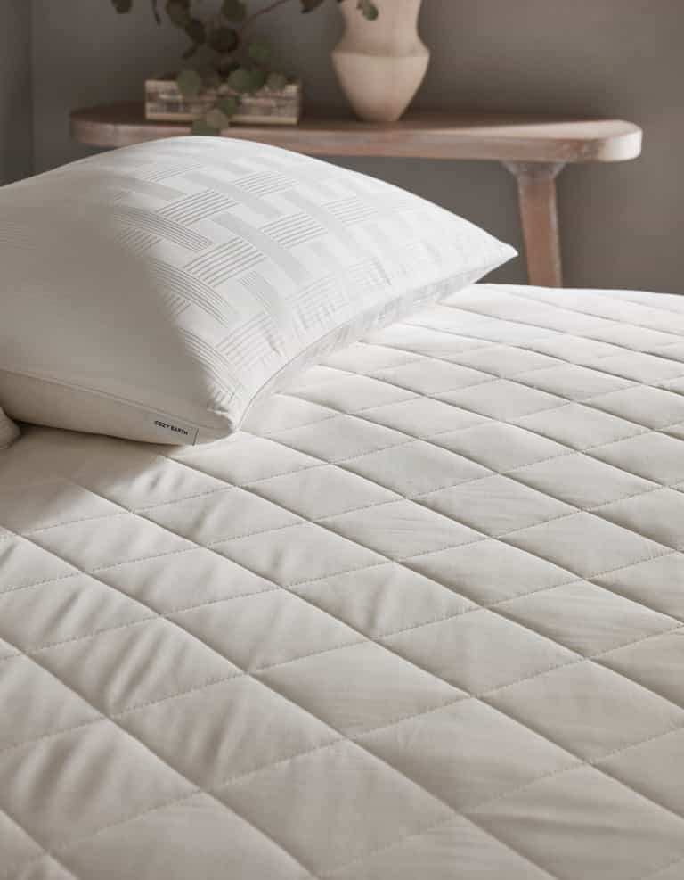 A neatly made bed features the Cozy Earth Bamboo Mattress Pad and a white pillow with a geometric pattern. In the background, a wooden table holds a ceramic vase with greenery.