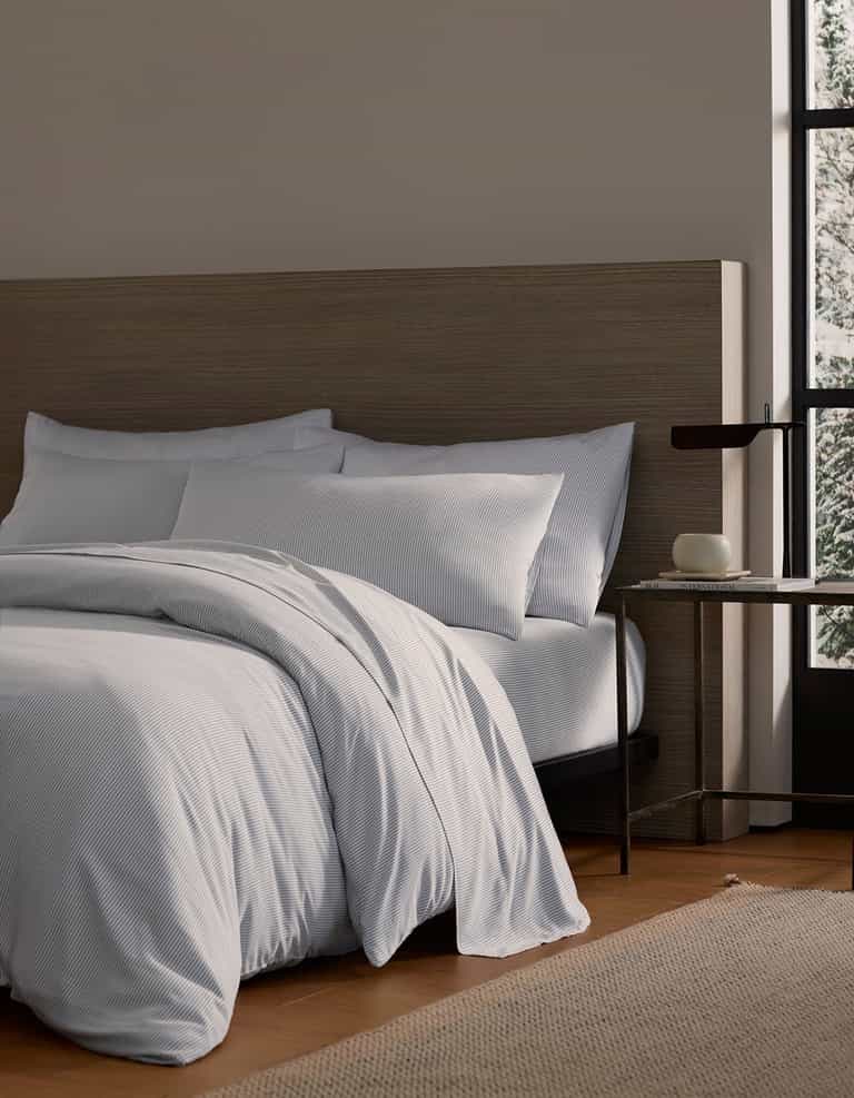 A Bamboo Duvet Cover by Cozy Earth in white covers a cozy bed with pillows against a wooden headboard. A small side table with a dark lamp and books sits nearby, while a large window reveals a snowy outdoor view.|Color:Charcoal Pinstripe