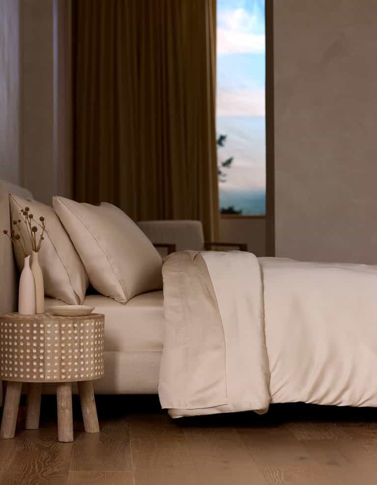 A neatly made bed with the Cozy Earth Bamboo Sheet Set in cream sits next to a round wooden nightstand holding two vases of dried flowers, as soft natural light streams through a window with beige curtains.|Color:Creme