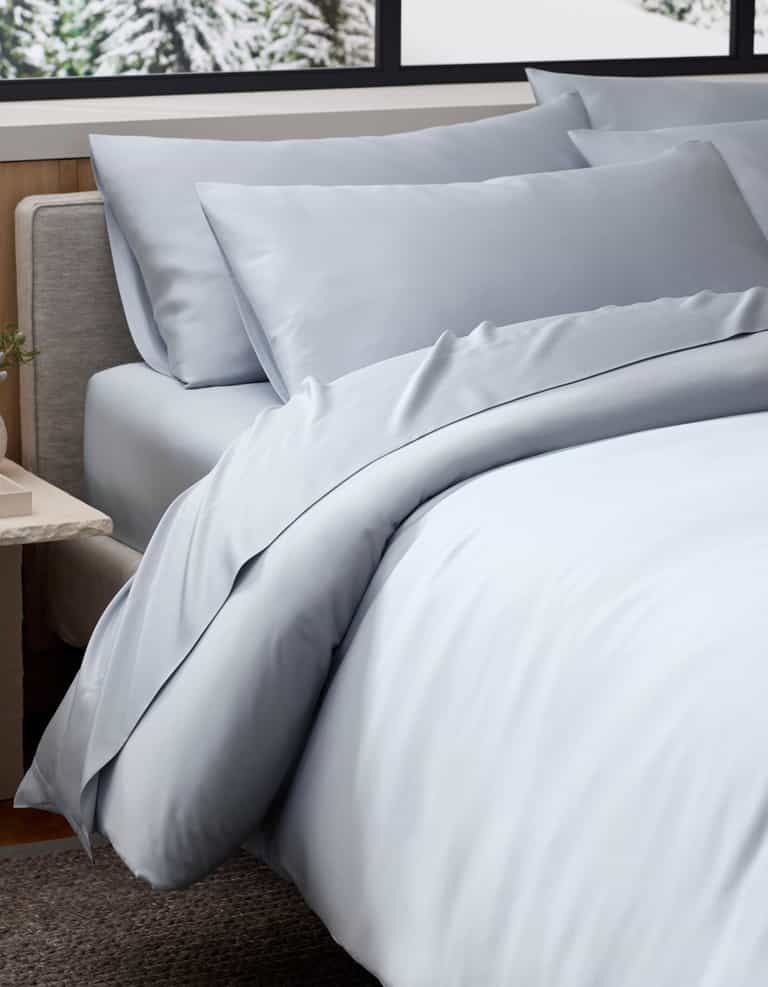 A neatly made bed features Cozy Earth's Bamboo Duvet Cover in light blue, with matching sheets and pillowcases, set beside a beige upholstered headboard and a bedside table, by a window overlooking a snowy landscape.