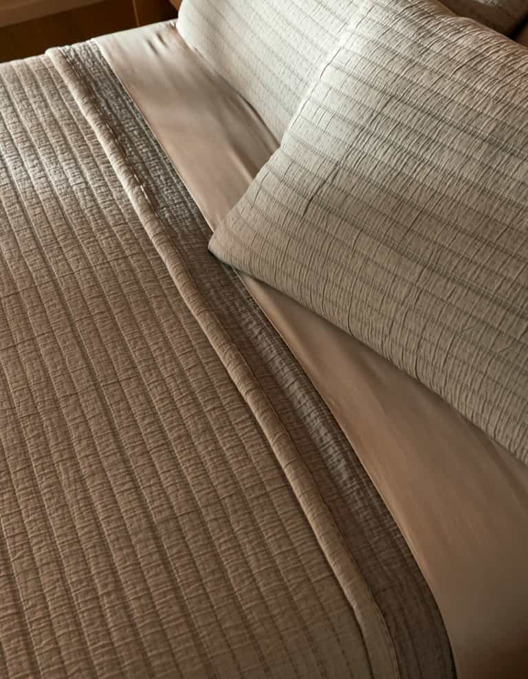 Close-up of a neatly made bed featuring the Cozy Earth Baja Matelasse Coverlet in light gray with matching pillows, accented by a smooth white sheet underneath. The bedding looks clean and inviting.|Color:Alabaster/Oceanside