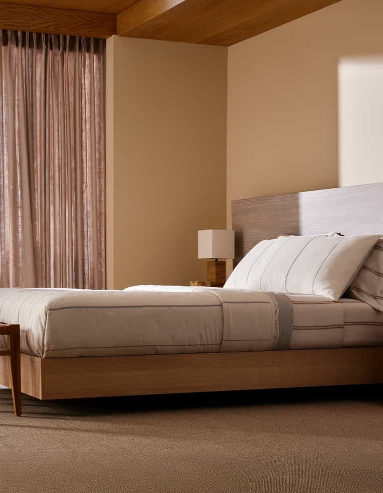 A modern bedroom features Cozy Earth’s Baja Quilt Bundle on a wooden platform bed, paired with a matching nightstand and lamp. Sheer curtains let in soft natural light, fostering a calm, minimalistic vibe.|Color:Coal Stripe/Striped Alabaster