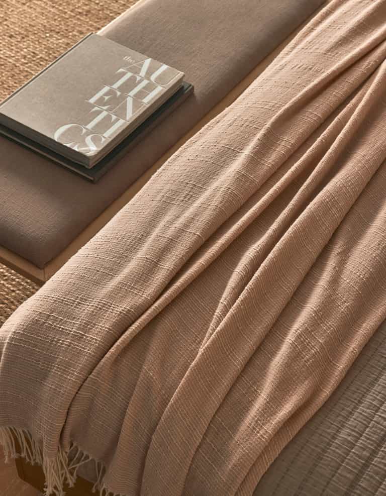 The Baja Tassel Throw by Cozy Earth, in beige with a textured finish, is draped over a neutral-toned bench beside two stacked books (one titled "AUTHENTICS"), creating a cozy, minimalistic look.|Color:Himalayan Salt