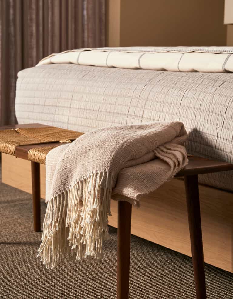 The Cozy Earth Baja Tassel Throw, a beige woven blanket with fringe, is draped over a wooden bench at the foot of a bed with a textured light bedspread, complementing the room's warm neutral tones and soft lighting.|Color:Himalayan Salt
