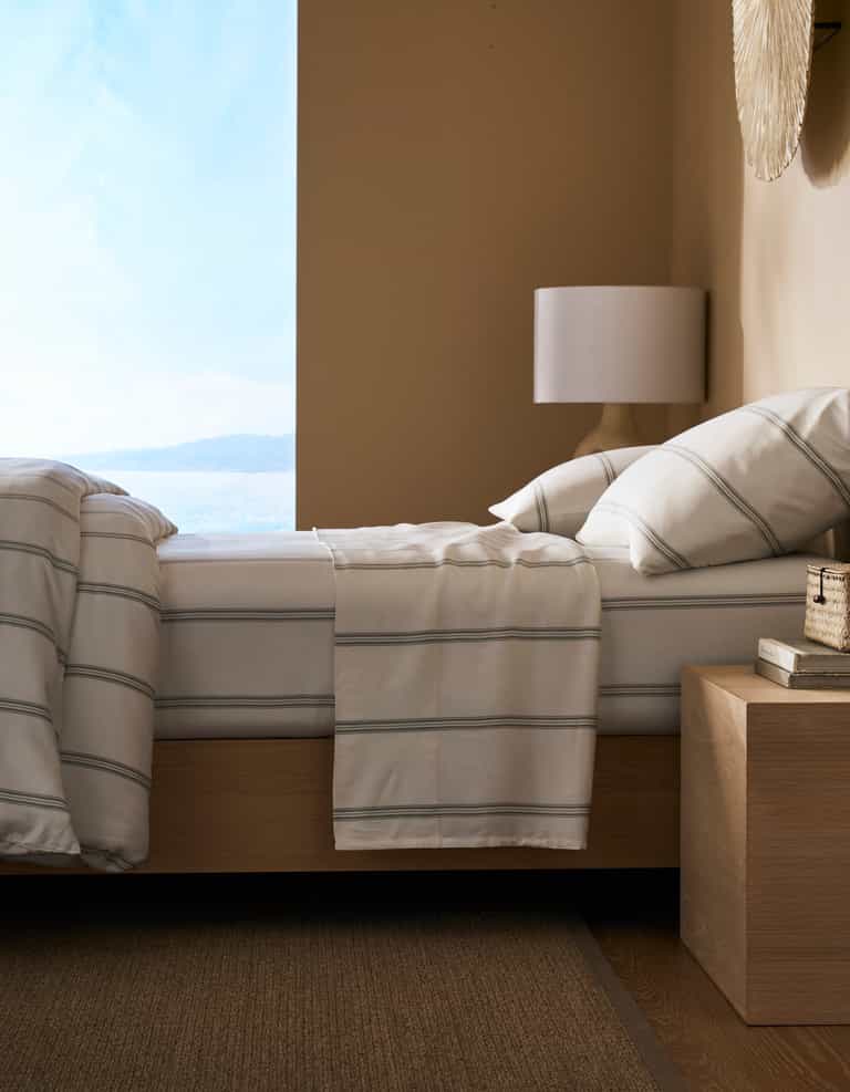 A modern bedroom featuring a neatly made bed with Cozy Earth’s Bamboo Sheet Set, a wooden nightstand with books and decor, a white lamp, and a large window offering views of water and sky.|Color:Baja Stripe in Laurel