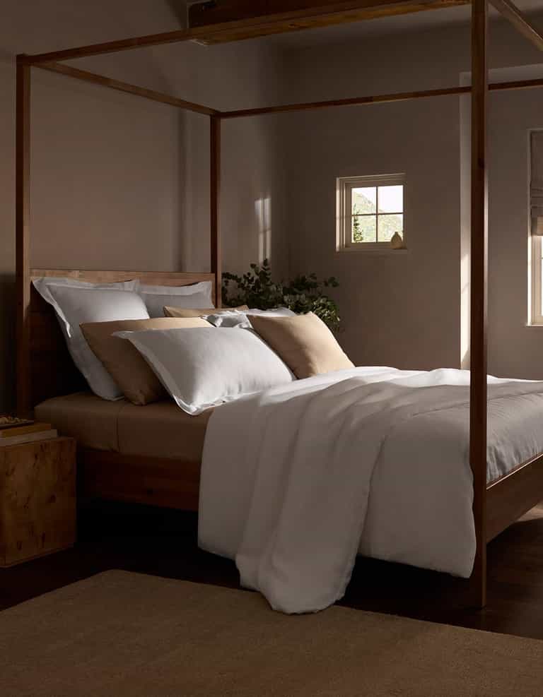 A cozy bedroom features a wooden four-poster bed with a Cozy Earth Linen Duvet Cover, soft light streaming through small windows, and a plant on the nightstand, creating a calm and inviting atmosphere.|Color:White