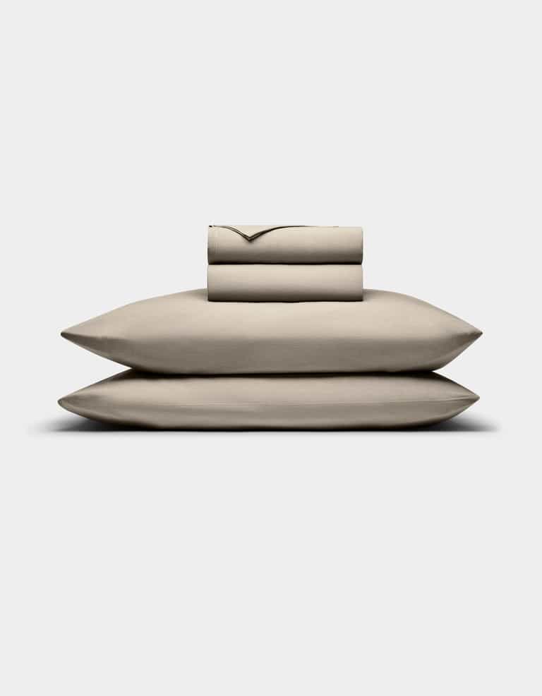 The Bamboo Jersey Sheet Set by Cozy Earth is displayed on a plain white background, featuring a neatly folded beige set with two stacked pillowcases, a fitted sheet, and a flat sheet.|Color:Driftwood
