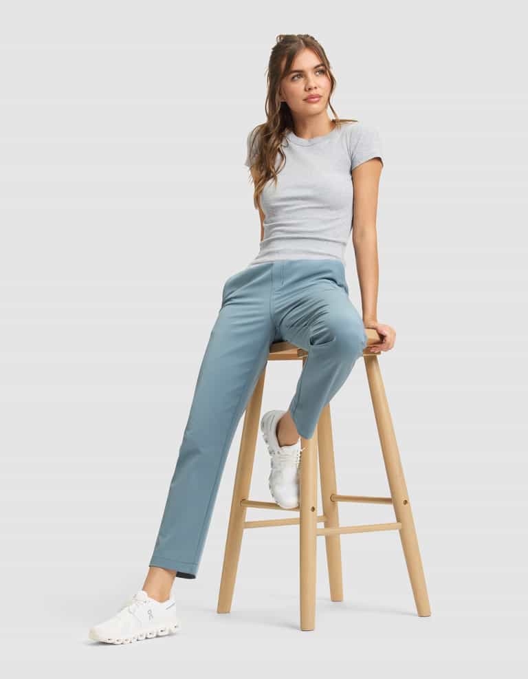 A woman in a grey t-shirt and white sneakers wears Cozy Earth's Women's Always Cropped Pant in light blue, sitting casually on a wooden stool against a plain, light grey background. |Color:Oceanside